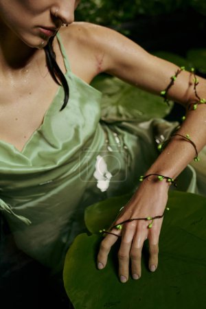 A woman in a green dress poses with a vine bracelet on a lily pad in a swamp.