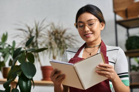 A young Asian woman in an apron is reviewing her notes while working in her plant shop.