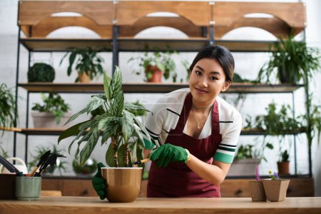A smiling Asian woman in an apron works in her plant store, tending to a vibrant green plant.