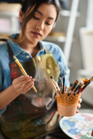 Eine junge asiatische Künstlerin mit Schürze hält einen Pinsel in der Hand und begutachtet verschiedene Pinsel in ihrer Werkstatt.
