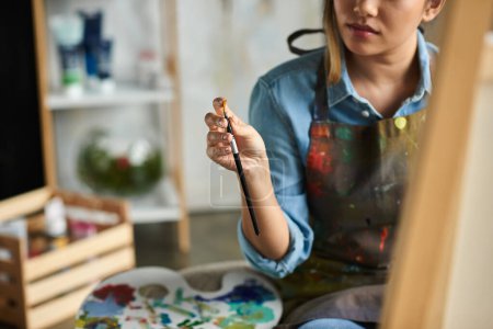 A young Asian woman, wearing an apron, is seen holding a paintbrush, focused on her artwork in her workshop.