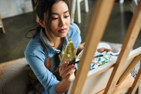 Eine junge asiatische Künstlerin mit Schürze malt in ihrer Werkstatt auf eine Leinwand.