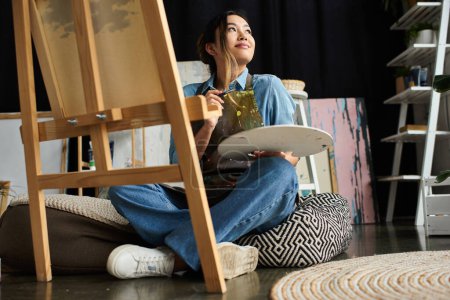 Eine junge asiatische Künstlerin sitzt in ihrer Werkstatt und hält eine Palette in der Hand, bereit, ein neues Meisterwerk zu schaffen.