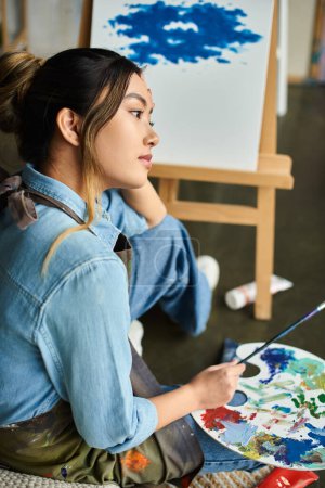 Woman in blue apron holds paintbrush in workshop, lost in thought over her painting.