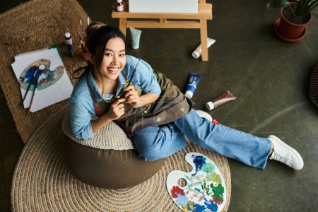 A young Asian woman, dressed in an apron, sits comfortably on a bean bag chair in her art studio, holding a paintbrush and smiling.