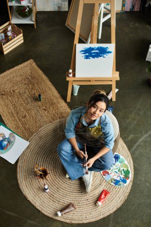 An Asian artist sits in her workshop, wearing an apron, and paints with brushes and paints.
