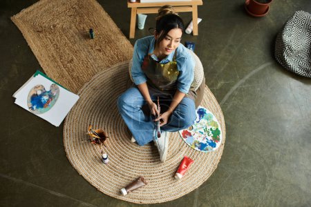A young Asian woman in an apron sits on a round rug, holding paintbrushes and reflecting on her artwork.