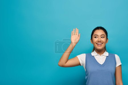 A young Asian woman in smart casual attire smiles and waves her hand against a blue background.