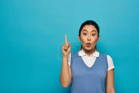 A young Asian woman in smart casual attire looks surprised with a raised finger, appearing to have just had a brilliant idea.