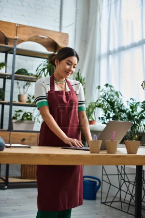 A beautiful Asian woman wearing an apron works on a laptop in her plant shop, surrounded by greenery.