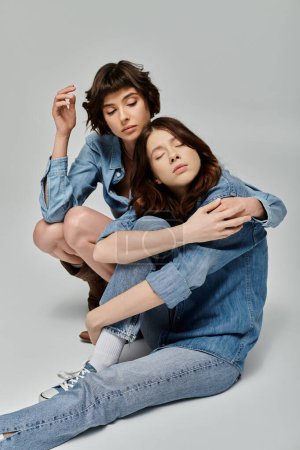 A young lesbian couple in stylish denim attire poses against a grey background.