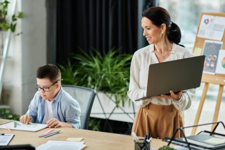 Une mère travaille sur son ordinateur portable dans son bureau, tandis que son fils trisomique est assis à une table de dessin.