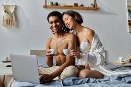 A young multicultural couple enjoys morning coffee and a laptop in their bedroom.