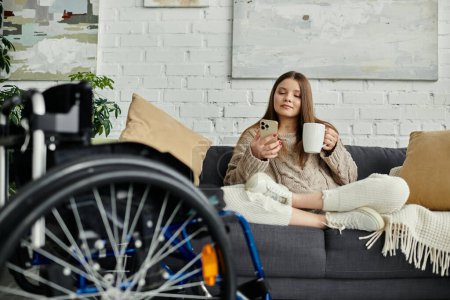 Eine junge Frau im Rollstuhl entspannt auf ihrem Sofa, genießt eine Tasse Kaffee und surft auf ihrem Handy..