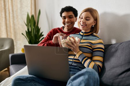 A happy multicultural couple enjoys a movie night on a couch in their modern apartment, sharing a bowl of popcorn while watching a laptop.