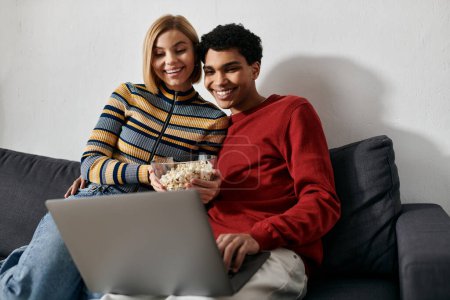 Ein fröhliches, multikulturelles Paar genießt einen gemütlichen Abend zusammen, schaut etwas auf seinem Laptop und teilt sich eine Schüssel Popcorn.