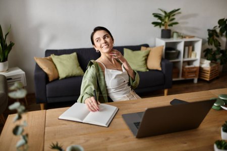 Une femme est assise à un bureau dans son bureau à la maison, écrivant dans un carnet tandis qu'un ordinateur portable est assis à côté d'elle.