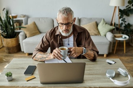 A mature gay man with grey hair and tattoos works from home, enjoying a cup of coffee.
