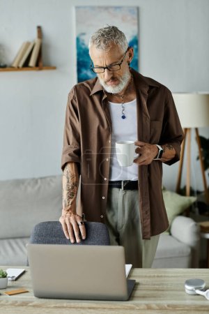 Un hombre gay maduro con tatuajes y cabello gris, trabajando remotamente desde casa, disfruta de una taza de café mientras trabaja en su computadora portátil.
