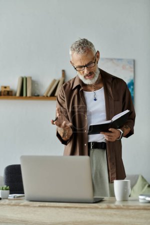 Ein reifer schwuler Mann mit grauen Haaren und Tätowierungen arbeitet von seinem Home Office aus mit Laptop und Notizbuch.