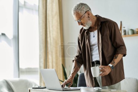A mature, tattooed man with grey hair works on his laptop while holding a cup of coffee.