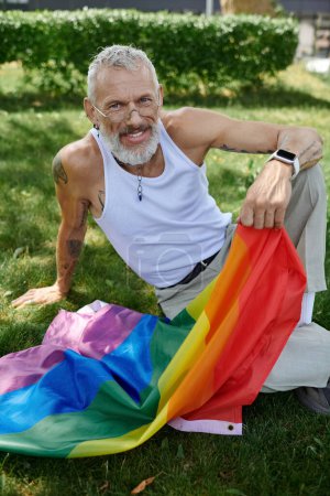 Ein reifer schwuler Mann mit Tätowierungen und grauem Bart lächelt, während er eine Regenbogenfahne in einem Park hält.