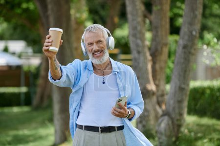 Ein älterer bärtiger Mann mit Tätowierungen genießt Kaffee, lächelt und hört Musik mit Kopfhörern.