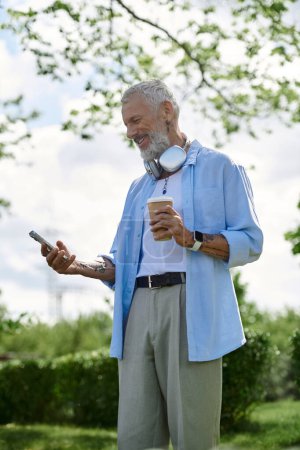 Ein reifer schwuler Mann mit Tätowierungen und grauem Bart sitzt auf dem Rasen in einem Park und genießt einen sonnigen Nachmittag.