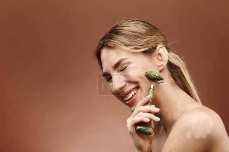 A young woman with vitiligo smiles as she uses a jade roller to massage her face, showcasing her skincare routine.