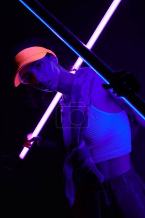 A young woman poses under vibrant neon lights, her orange cap casting a shadow across her face.
