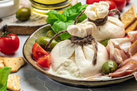 Italian tomatoes and burrata cheese salad served with cherry tomatoes, fresh basil leaves, prosciutto ham and olives on a light background. Restaurant menu top view.