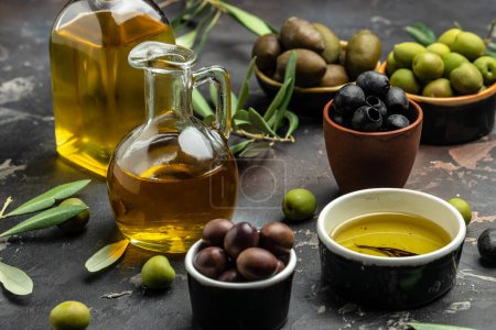 Foto de Aceite de oliva en dos recipientes de vidrio y recoger las bayas de aceitunas en cuencos sobre un fondo oscuro. - Imagen libre de derechos