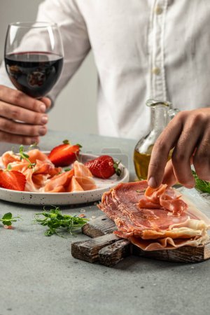 Photo for Hands holding a glass of wine and a wooden board with different kinds of cheese and ham, prosciutto, jamon salami, Antipasto Dinner or aperitivo party concept. - Royalty Free Image