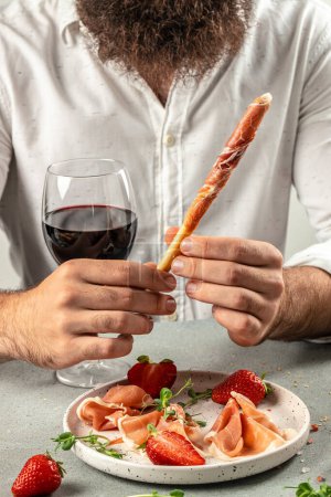 Photo for Hands holding a glass of wine and a wooden board with different kinds of cheese and ham, prosciutto, jamon salami, Antipasto Dinner or aperitivo party concept. - Royalty Free Image