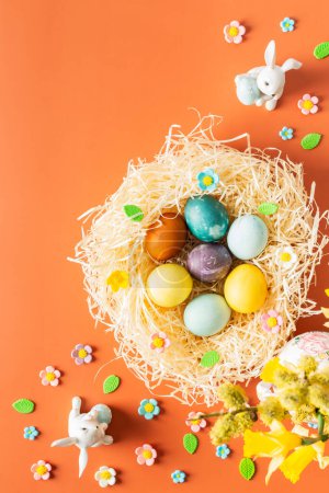 Top view of nest with natural dyes dyed Easter eggs, bouquet with yellow daffodils in vase and Easter bunnies on orange background