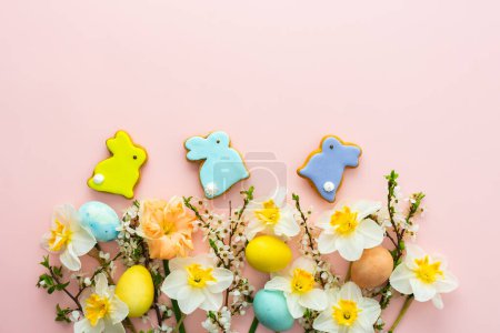 Fondo festivo con flores de primavera y huevos de colores naturales y conejitos de Pascua, narcisos blancos y ramas de flor de cerezo sobre un fondo pastel rosa