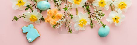 Banner festivo con flores de primavera y huevos de colores naturales y conejitos de Pascua, narcisos blancos y ramas de flor de cerezo sobre un fondo pastel rosa
