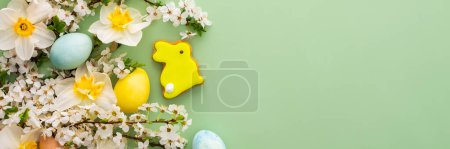 Banner festivo con flores de primavera y huevos de colores naturales y conejitos de Pascua, narcisos blancos y ramas de flor de cerezo sobre un fondo verde pastel