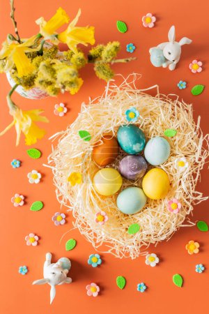 Vue du dessus du nid avec des colorants naturels teints oeufs de Pâques, bouquet avec jonquilles jaunes dans un vase et lapins de Pâques sur fond orange