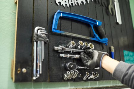 Téléchargez les photos : Main d'une personne méconnaissable portant un gant de protection saisissant un objet sur une planche à outils dans le mur d'un atelier de réparation de vélos. - en image libre de droit