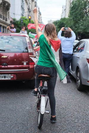Foto de Buenos Aires, Argentina, 16 de febrero de 2024: Mujer en bicicleta con una bandera palestina en la espalda y un puño levantado, en una protesta de caravana en solidaridad con Palestina y contra el ataque genocida de Israel. - Imagen libre de derechos