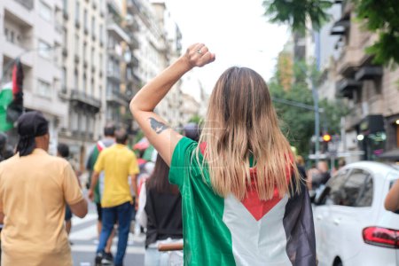 Foto de Buenos Aires, Argentina, 16 de febrero de 2024: Mujer con una bandera palestina en la espalda y un puño levantado durante una manifestación en solidaridad con Palestina y contra el ataque genocida de Israel. - Imagen libre de derechos