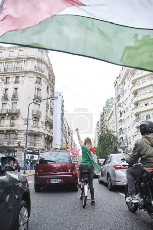 Foto de Buenos Aires, Argentina, 16 de febrero de 2024: Mujer en bicicleta con una bandera palestina en la espalda y un puño levantado, en una protesta de caravana en solidaridad con Palestina y contra el ataque genocida de Israel. - Imagen libre de derechos