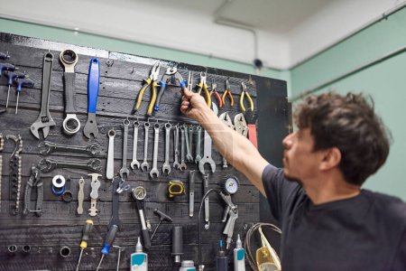 Téléchargez les photos : Un réparateur hispanique saisit un coupe-câble d'une planche à outils dans le mur d'un atelier de réparation de bicyclettes. De vraies personnes au travail. - en image libre de droit