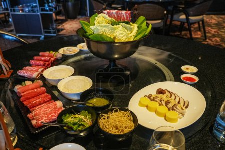 A hot pot meal setup with various raw meats, vegetables, noodles, and sauces on a table.