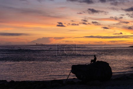 Une figure solitaire est assise sur un rocher au bord de la mer, méditant à la fin de la journée avec un magnifique affichage des couleurs du coucher de soleil peignant le ciel et réfléchissant sur l'eau