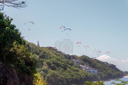 A group of colorful paragliders flies high above the cliffside coast of Bali, showcasing the thrill of adventure sports against the stunning tropical landscape
