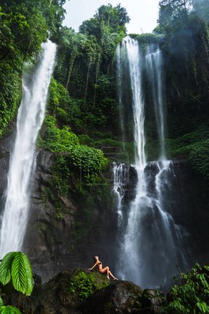 Woman lounging on a rock in front of a massive waterfall in a tropical forest.