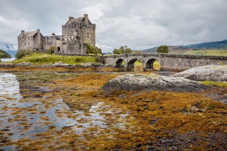 En la parte inferior del puente, sobre un cuerpo de agua, se puede ver el castillo de Eilean Donan. El castillo está rodeado de rocas y agua. Uno de los paisajes más bellos de Escocia. 