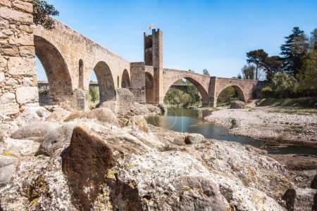 The medieval bridge of Besalu crosses the Fluvia river with a stone wall on each side. The bridge is old and has a rustic look.